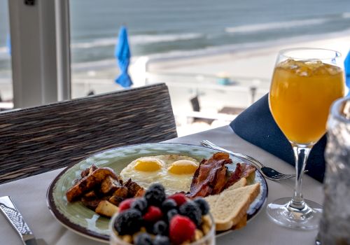 A breakfast plate with eggs, bacon, toast, potatoes, and berries, alongside a glass of iced tea, set on a table overlooking a beach.