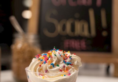 A cup of ice cream topped with whipped cream and sprinkles, with a blurred sign behind it saying 