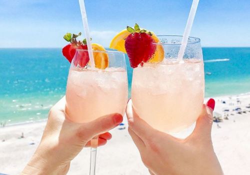Two hands holding glasses with pink beverages, garnished with strawberries and orange slices, in front of a beach and ocean view.